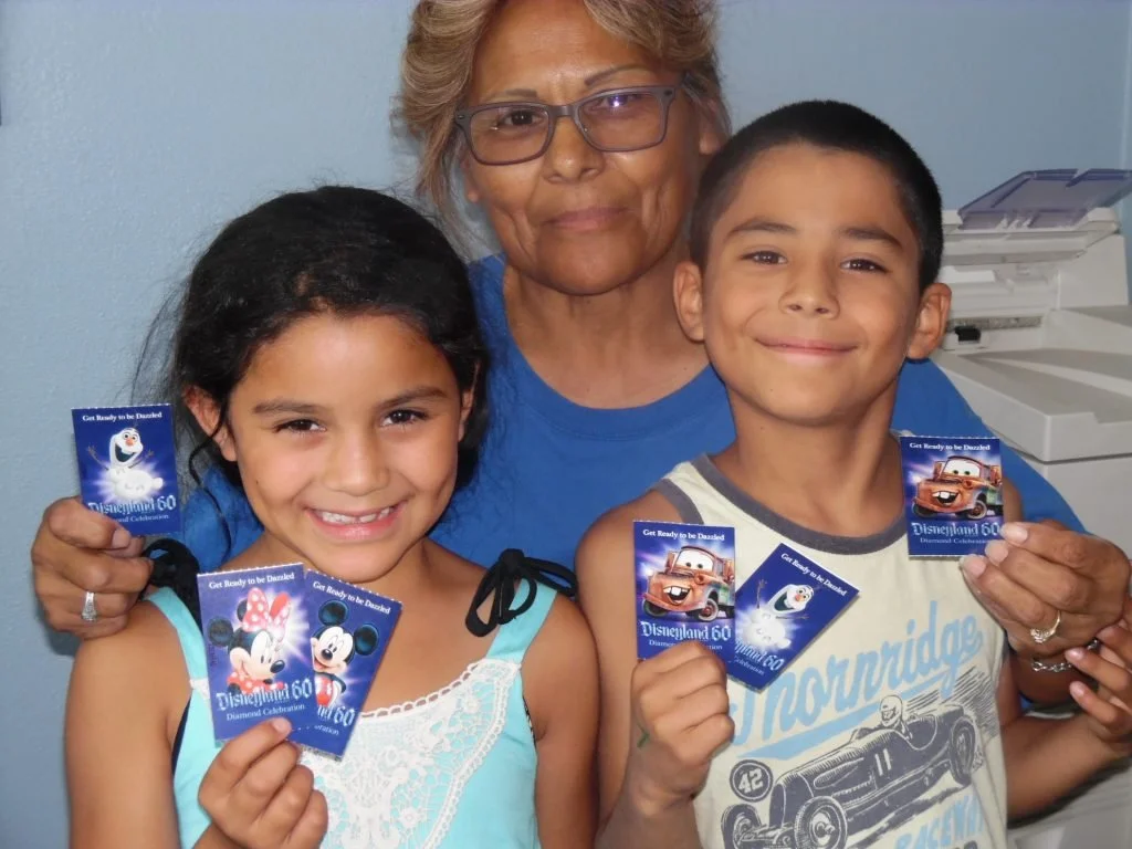 A woman and two children smiling and holding Disneyland 60th anniversary tickets inside a room with a copier in the background.