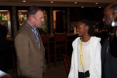 Two men and a woman having a conversation in a restaurant or bar setting, with wooden furnishings and dim lighting.