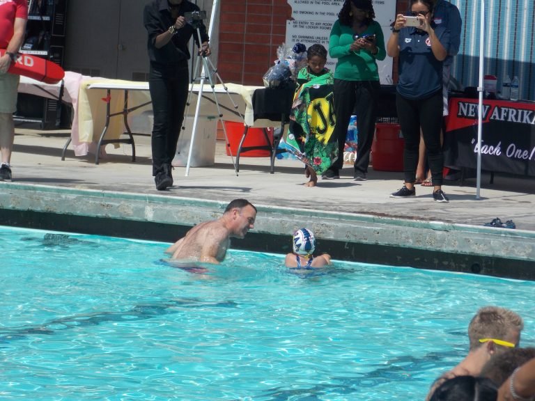 A man and a young girl in a swimming pool, with a group of people on the pool deck watching and taking photos.