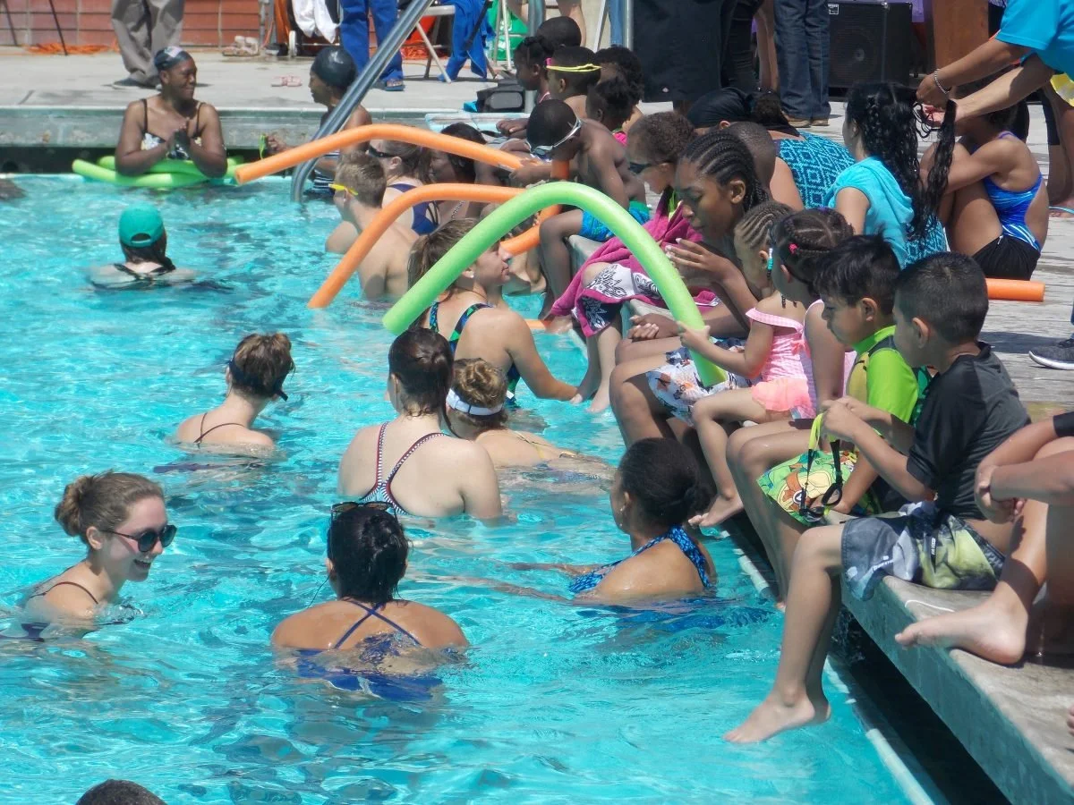 Children and adults gather at the edge of a swimming pool, with some in the water and others sitting on the poolside, participating in a swimming lesson or activity on a sunny day.