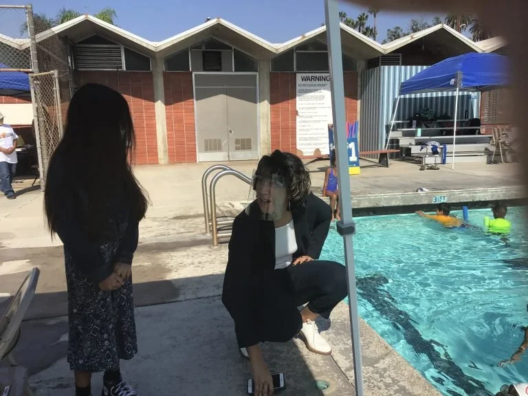 A woman crouching near the edge of a swimming pool, talking to a girl standing beside her. Two children are swimming in the pool, and there is a tent and some pool equipment in the background, along with a building.