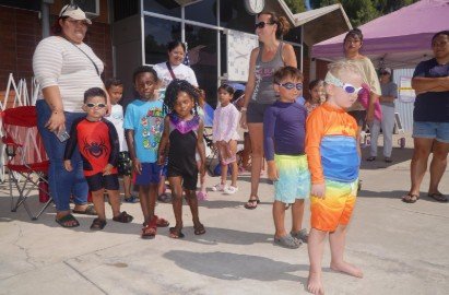Group of children and adults gathered outside, some children wearing colorful swimsuits and sunglasses, near a building with a purple canopy.