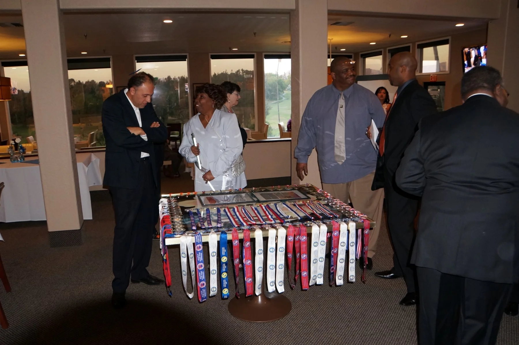 People gathered around a table with medals at an indoor event, with large windows revealing a golf course outside.