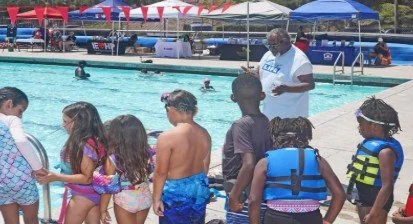Children and adults gathered at a swimming pool during a swimming safety or class event, with some children wearing life jackets.