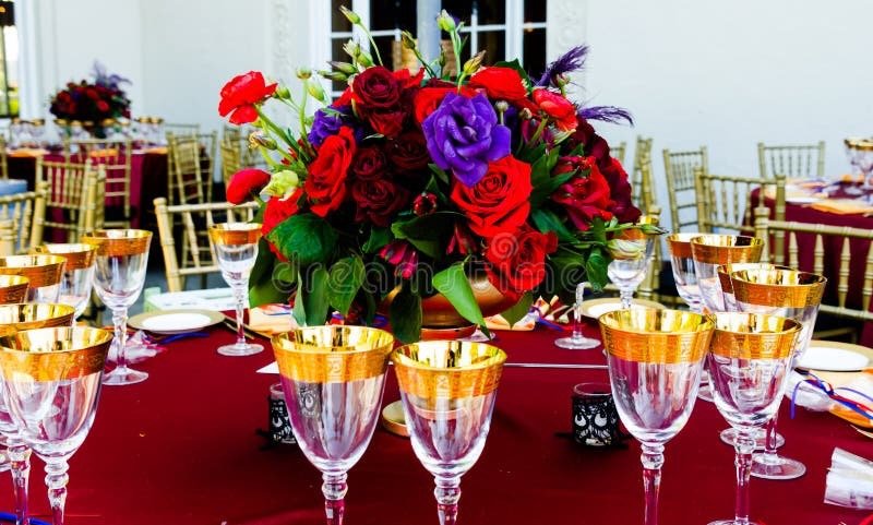 A table decorated with a large flower arrangement with red and purple flowers, surrounded by glasses with gold rims in a banquet hall.