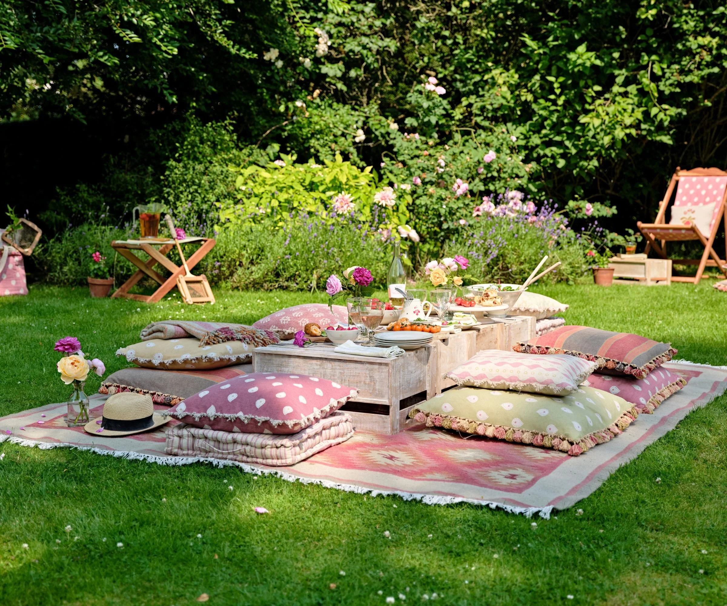 A picnic setup on a grassy lawn with colorful cushions on a rug, a low wooden table with food and drinks, flowers in vases, and garden chairs in the background surrounded by greenery.