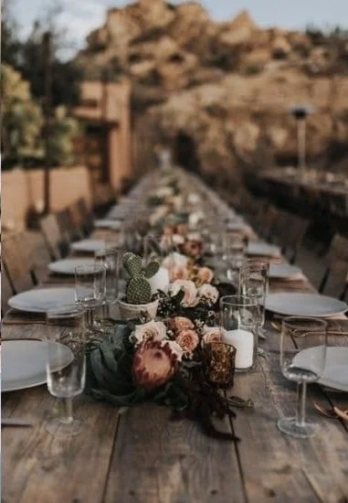 Long outdoor wooden dining table set for a meal, decorated with floral centerpieces and glassware, outdoors in a scenic area.