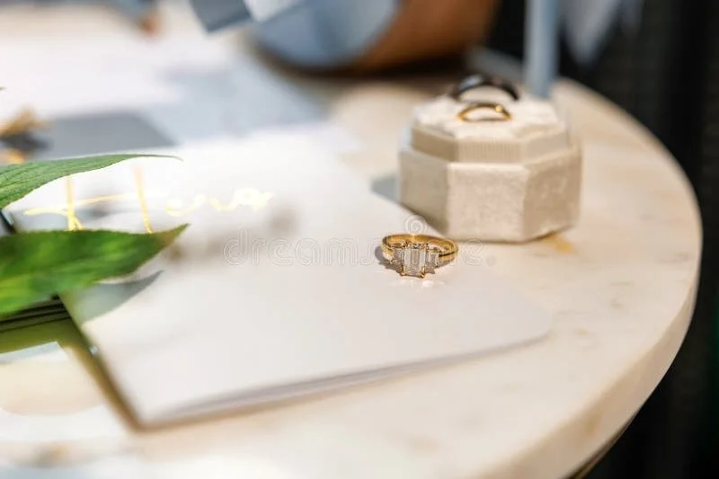 A gold ring with a rectangular diamond is placed on a white paper on a marble table, with a green leaf partially visible on the left and a small jewelry box in the background.
