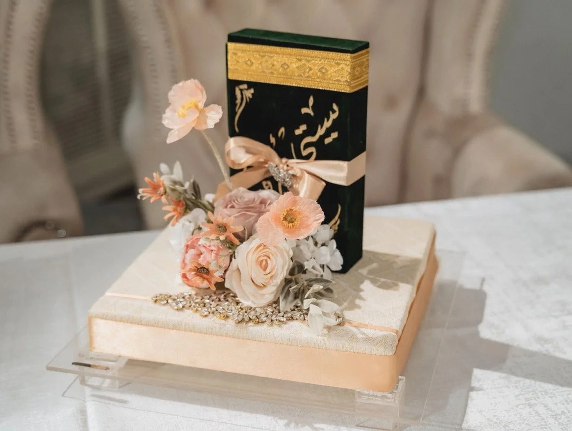 A wedding cake display featuring a black and gold rectangular box tied with a peach ribbon, decorated with a pink flower and surrounded by a floral arrangement with roses, poppies, and other flowers, placed on a white cake board with a pearl necklace and rhinestone embellishments.