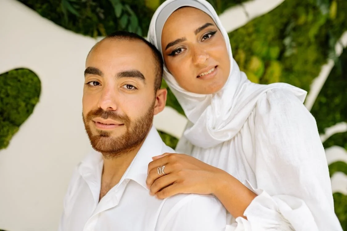 A woman wearing a white headscarf and white blouse sits beside a man with a beard and short hair, both dressed in white, posing together outdoors against a green leafy background.