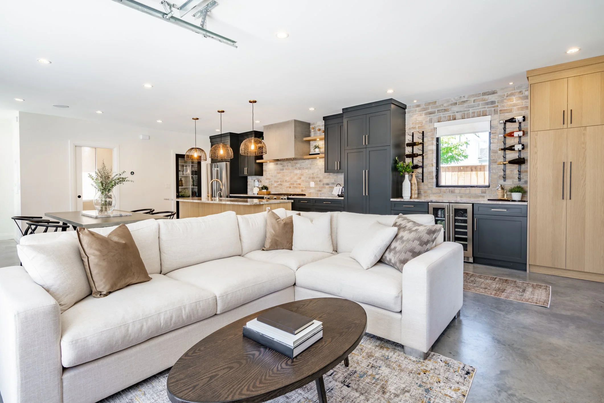 Open-concept living room and kitchen with white sectional sofa, dark cabinets, brick backsplash, pendant lights, and window with outdoor view.