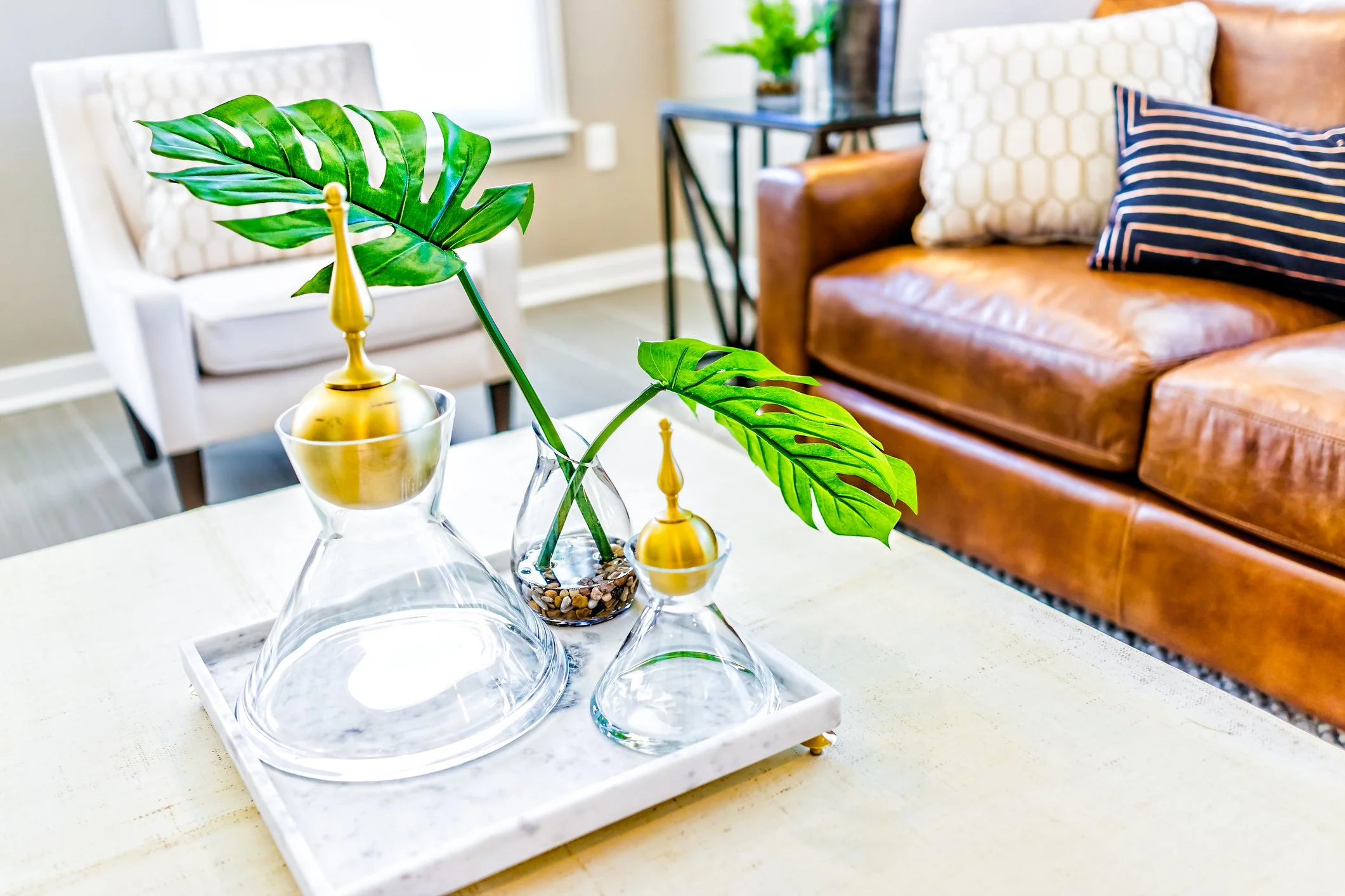 Living room with a white armchair, a brown leather sofa with pillows, a black side table with books, and a white tray with decorative glass vases and a large green monstera leaf.