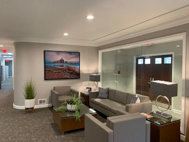 Modern medical lobby area with gray sofas, a glass coffee table decorated with potted plants, a side table with a lamp, framed cityscape artwork on the wall, and a large glass partition door.