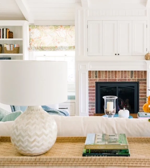 Living room with white walls, a brick fireplace, white built-in cabinets, a window with floral curtain, and a accent table with a white ceramic lamp and stacked books.