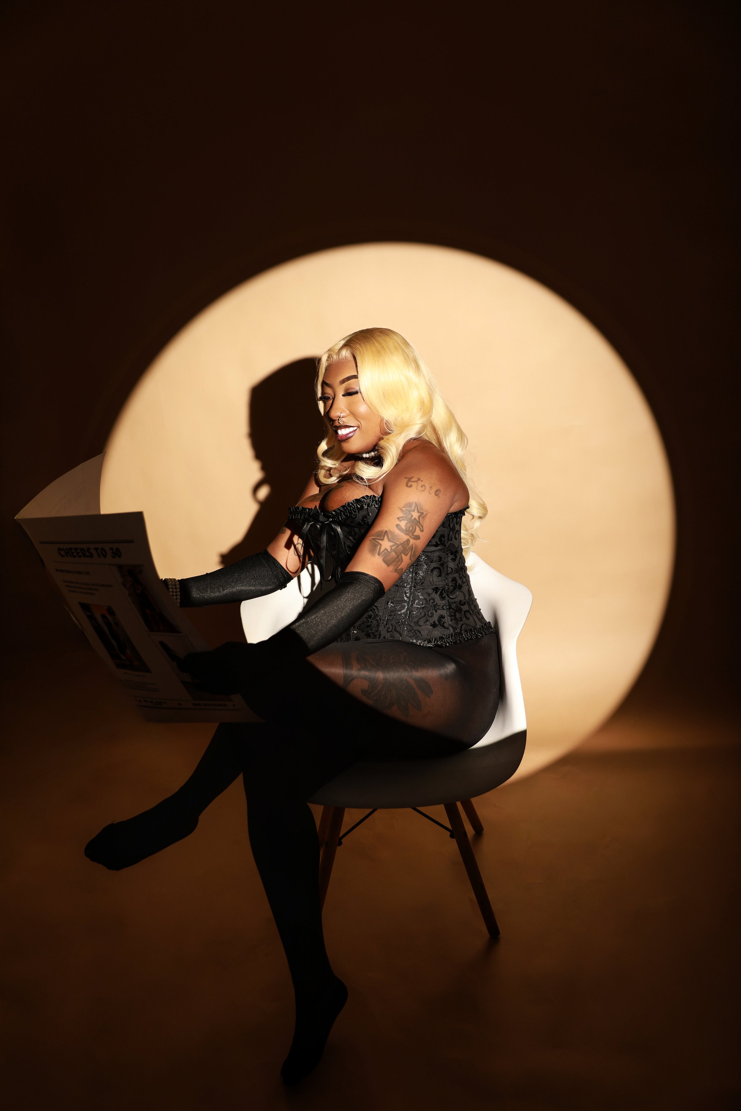 A woman with blonde wavy hair and tattoos on her arms, dressed in black lace lingerie and stockings, sitting on a white chair in a spotlighted setting, reading a newspaper and smiling.