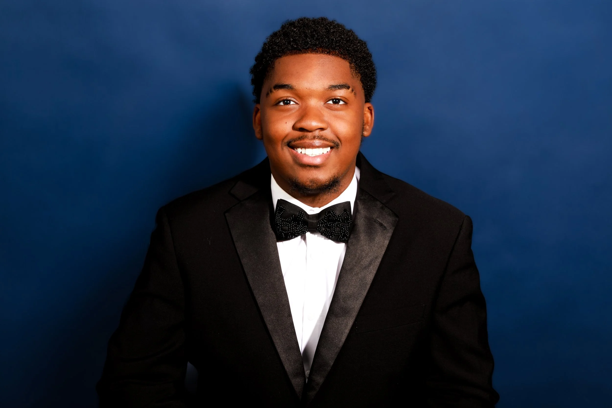 Young man in a tuxedo with a bow tie, smiling against a dark blue background.