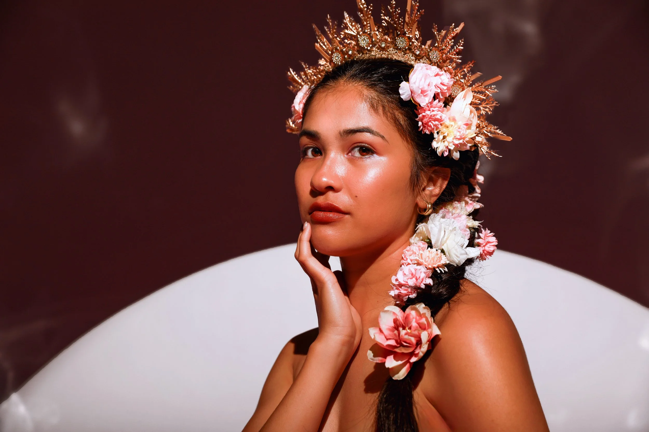 A woman with a floral crown and braid, touching her face gently, against a dark background.