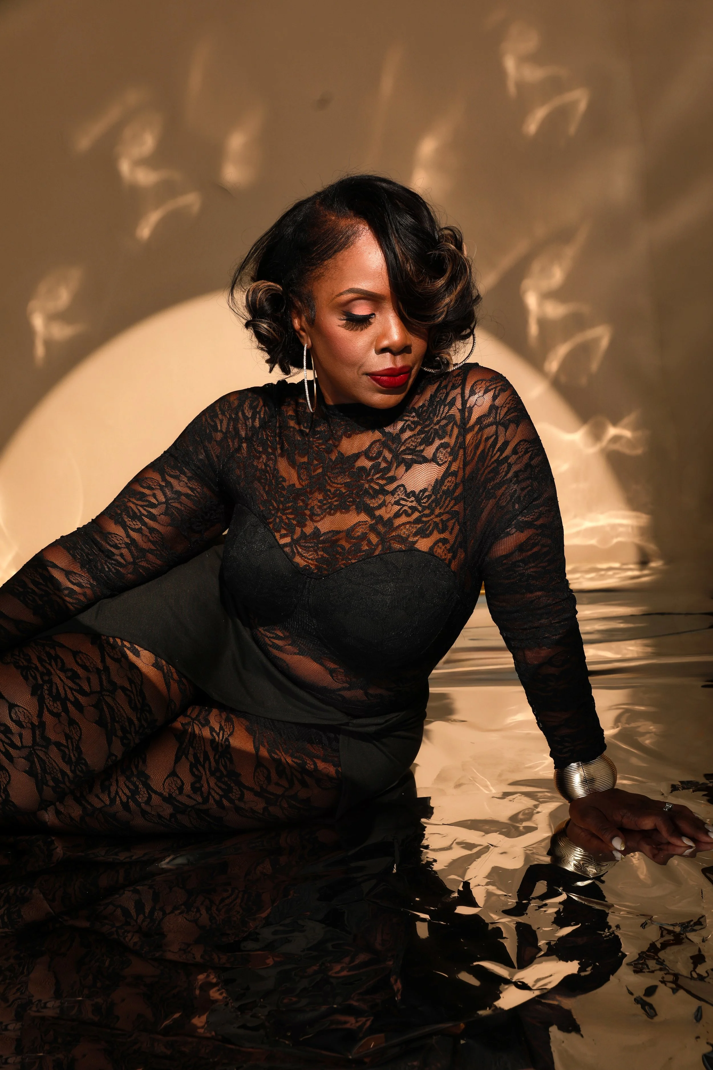 A woman with styled hair and red lipstick, dressed in black lace clothing, is sitting on a reflective surface with a dramatic spotlight behind her, creating an artistic shadow pattern on the wall.