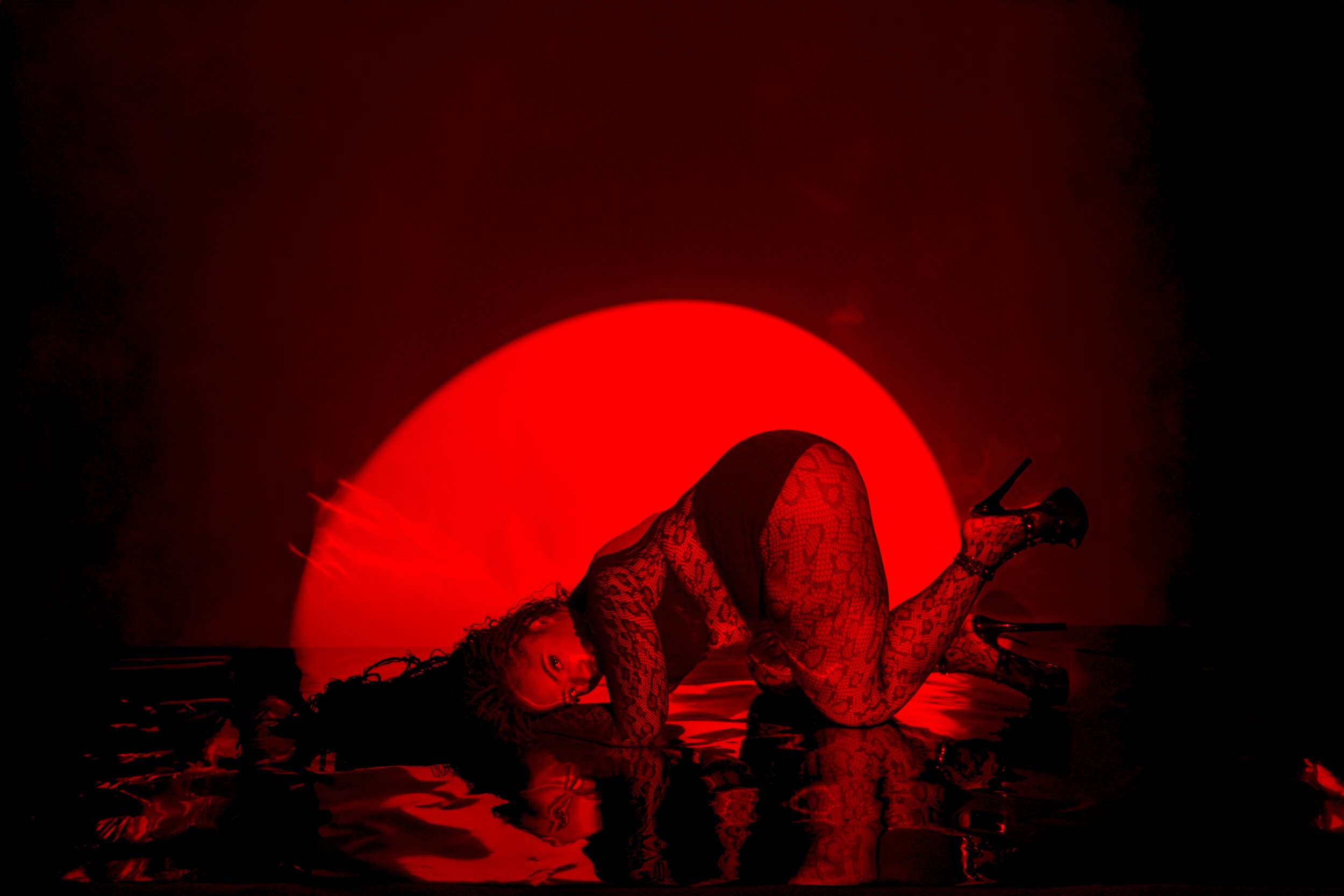 A woman in high heels, patterned stockings, and a lace dress is kneeling on the floor, arching her back with her head turned toward the camera, illuminated by a large red circle behind her in a dark room.
