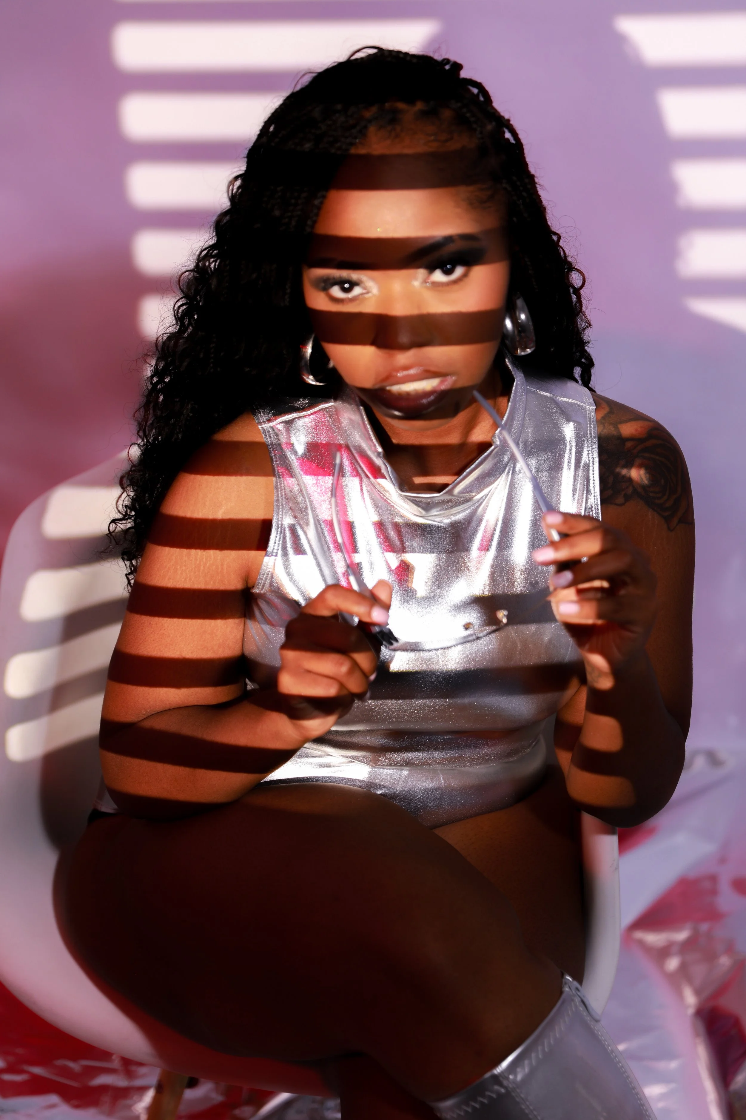 A woman with dark curly hair and dark lipstick is sitting in a white chair. She is wearing a shiny, metallic silver top and holding sunglasses. Light patterns create horizontal shadow lines across her face and body, casting a striped shadow effect.