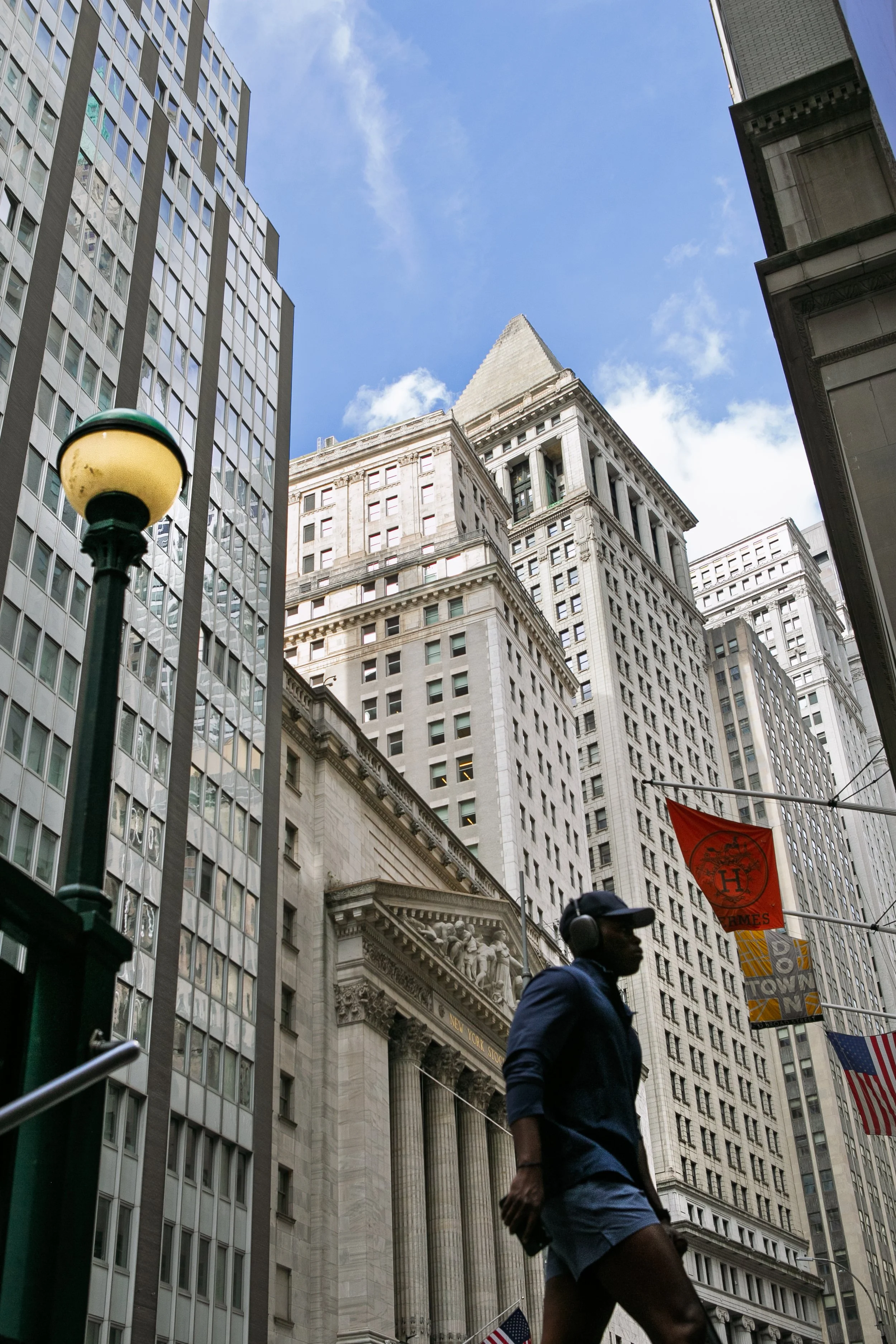 A person walking with headphones on in a city with tall buildings, including a historic building with columns and decorative sculptures, American flags, and street lamps, under a partly cloudy sky.
