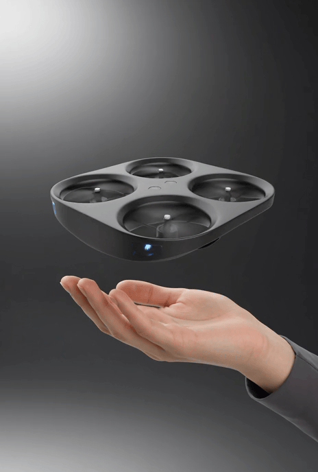 A hand holding out a square, four-compartment black drone with a sleek, modern design, floating above the palm against a dark background.