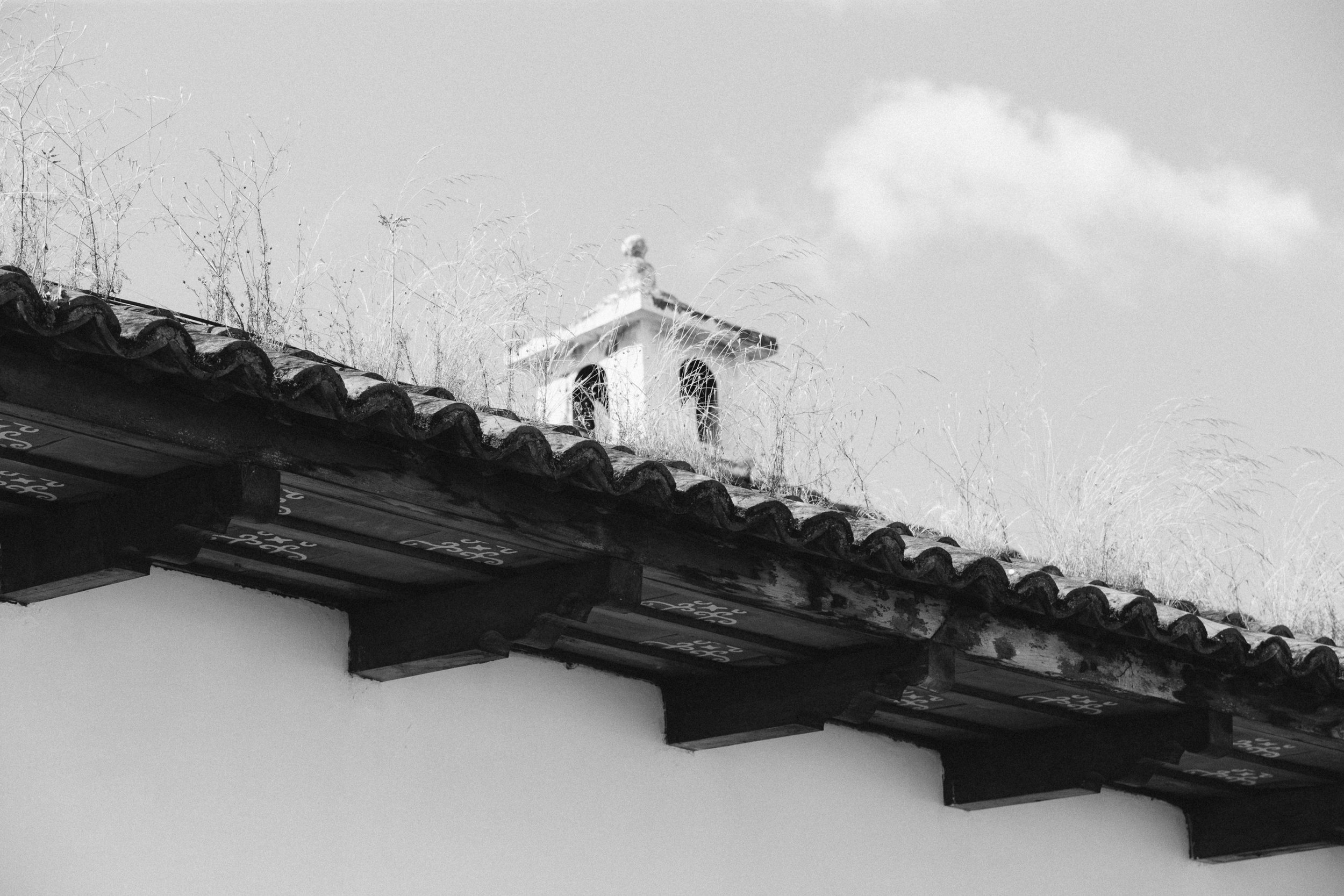 Black and white photograph of a tiled roof with a small decorative structure projecting from the top, surrounded by sparse dry grass and a partly cloudy sky.