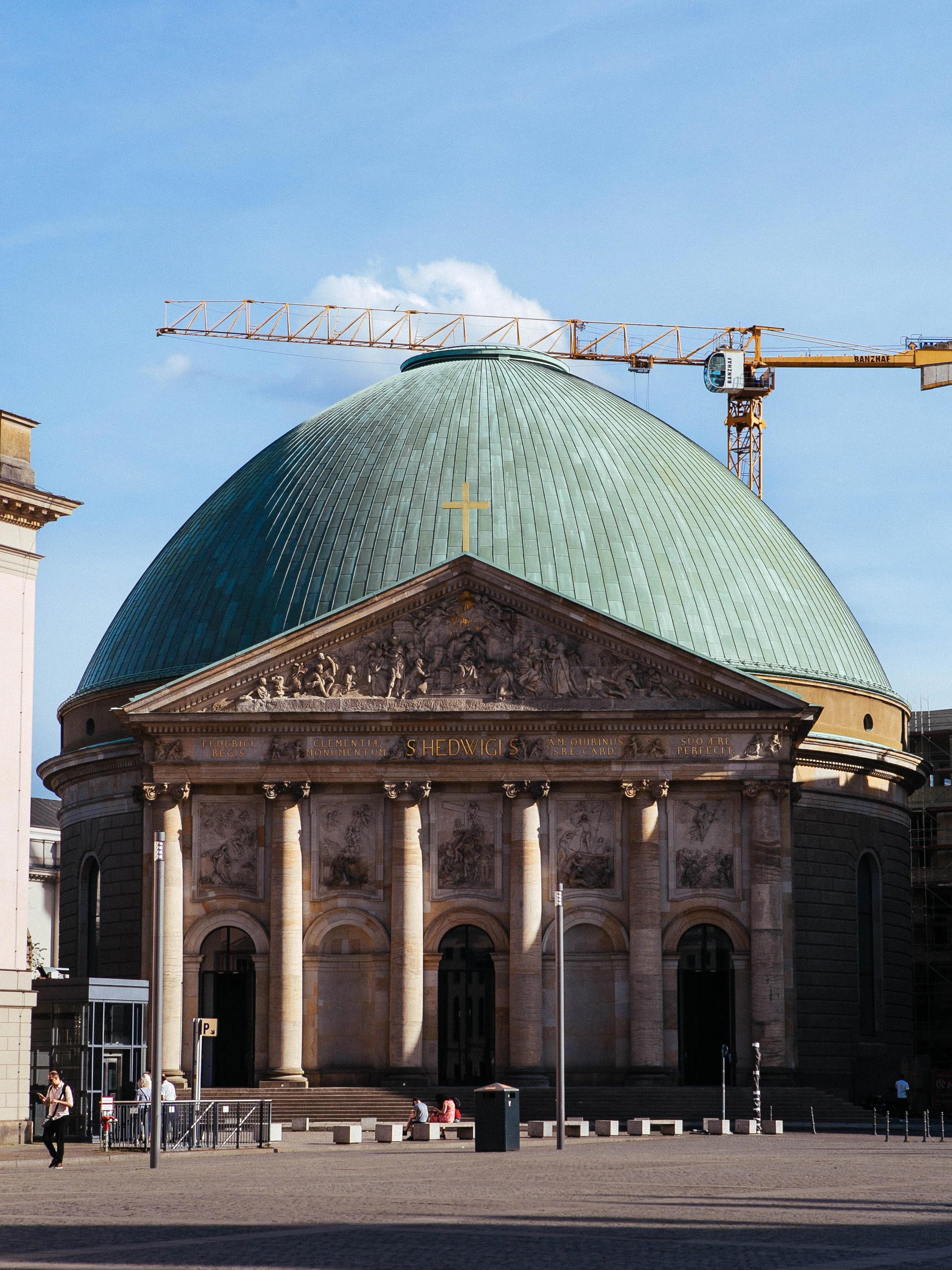 A church with a large green dome topped with a cross, with a construction crane in the background and people sitting or walking outside.