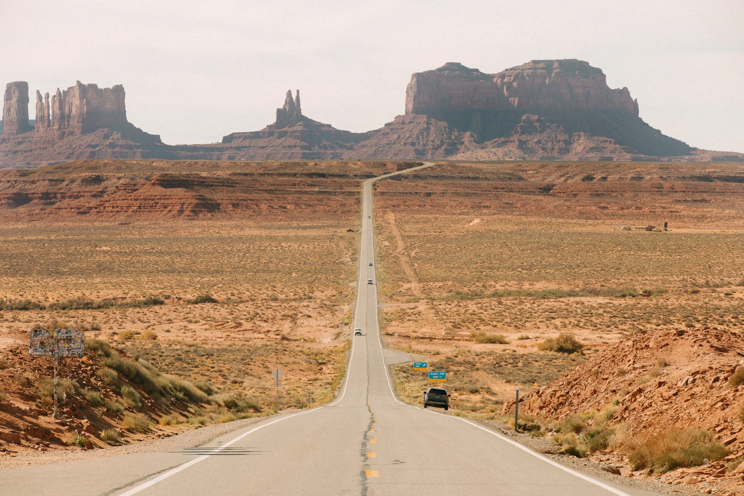 A long, straight road extending into the distance through a desert landscape with rock formations and mesas in the background.