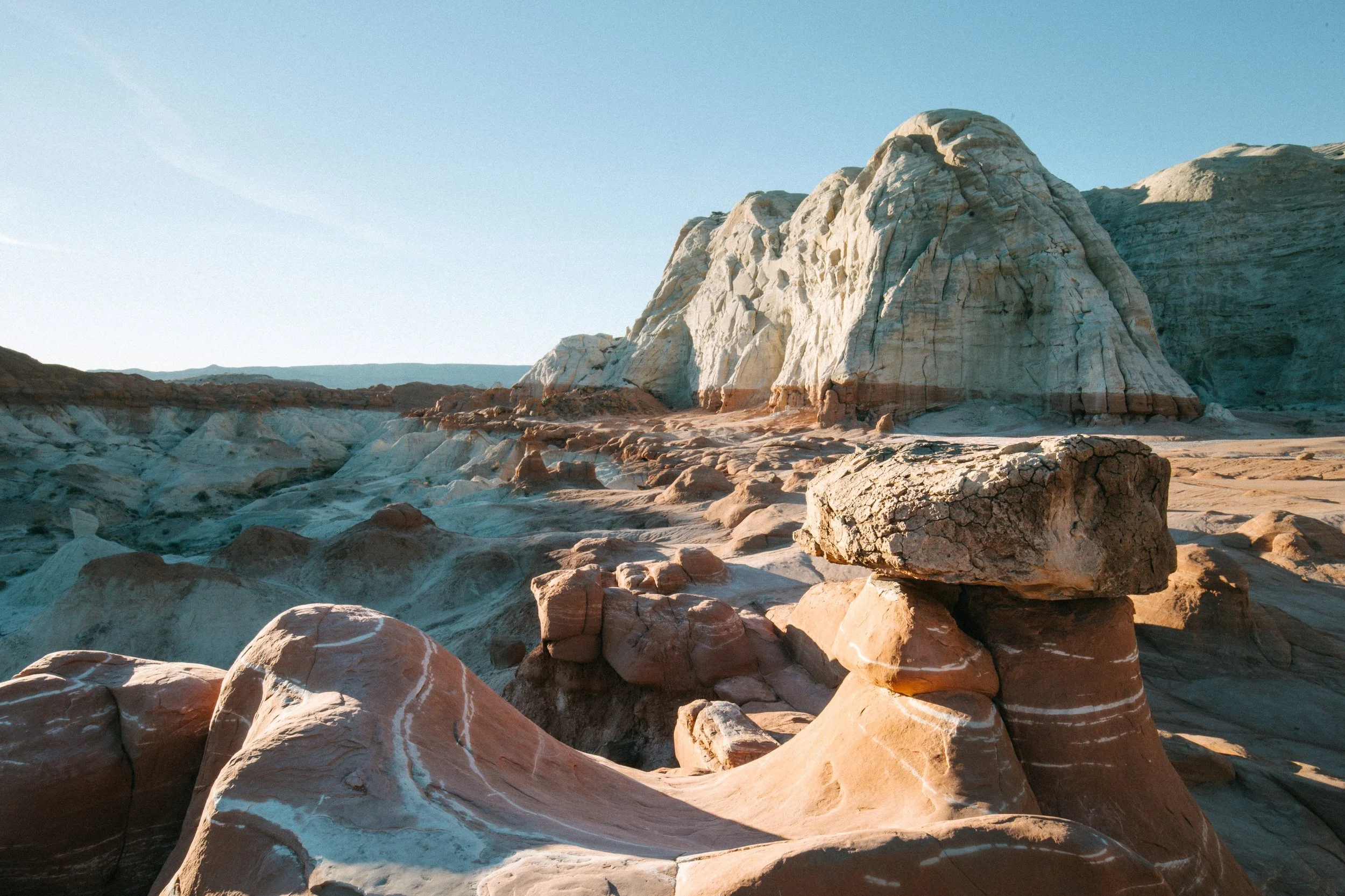Desert landscape with colorful rock formations and large white and reddish-brown sandstone cliffs in the background under a clear blue sky.