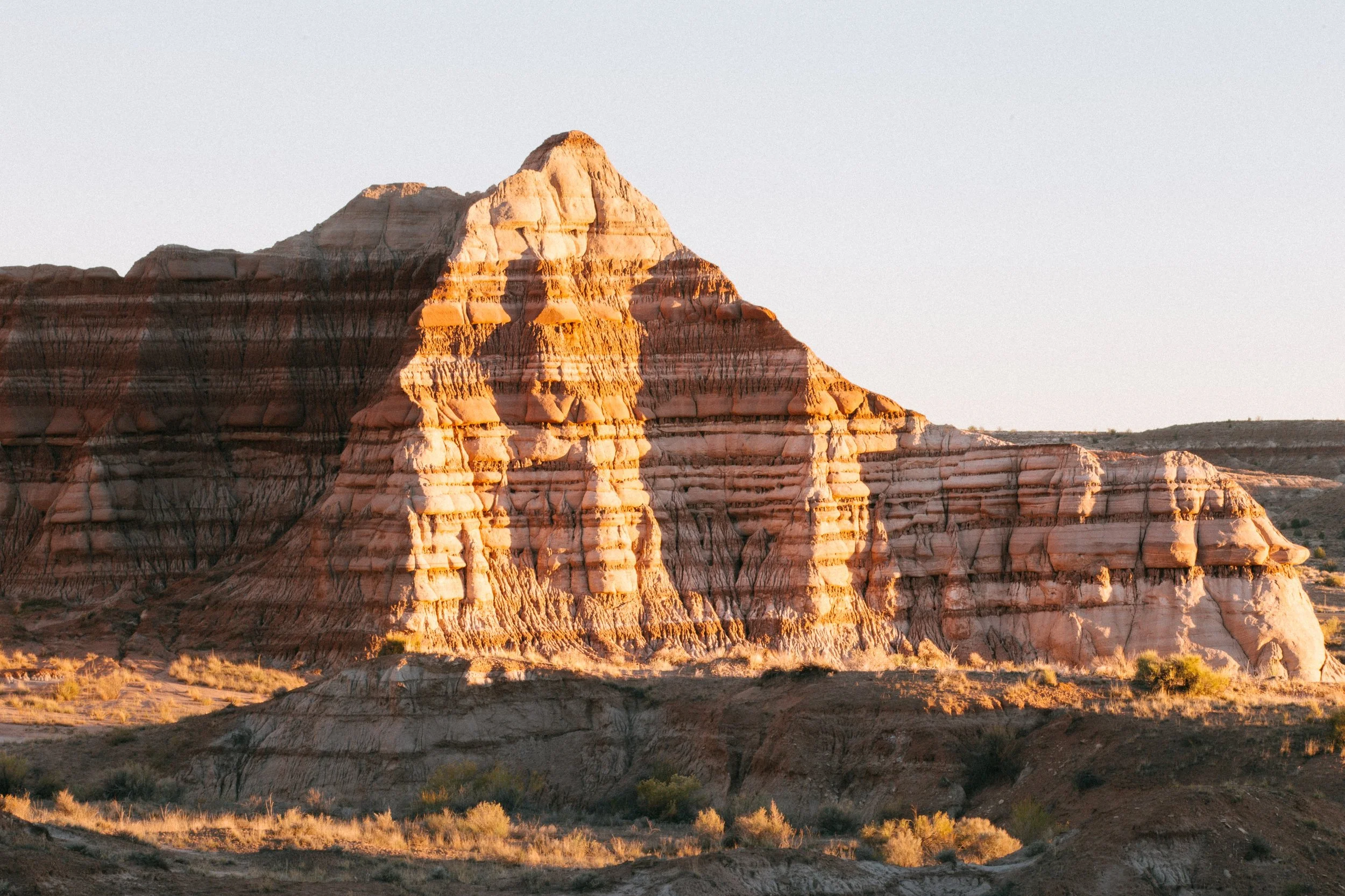 A large, layered desert rock formation with jagged peaks, illuminated by sunlight in a barren landscape.