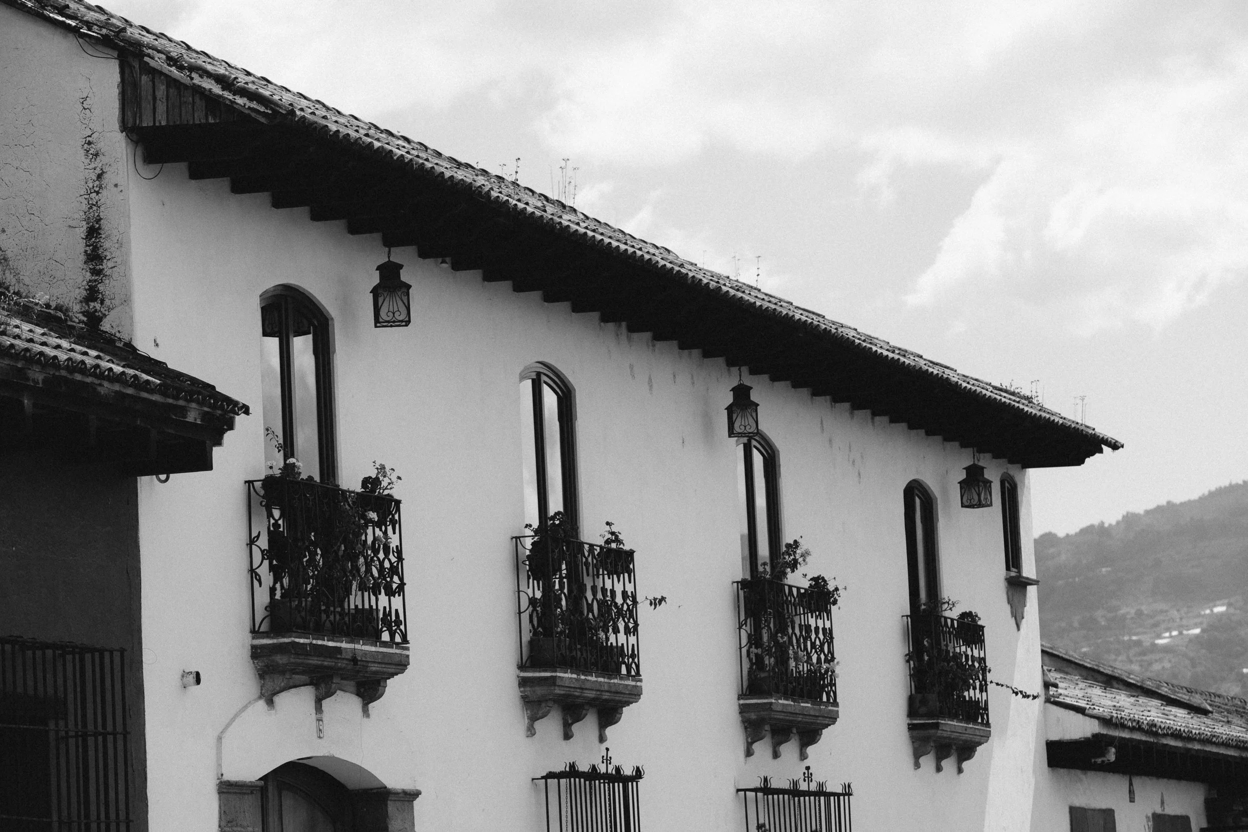 Black and white photo of an old building with arched windows, decorative balconies with plants, and lantern-style lights hanging from the roof, with mountains in the background.