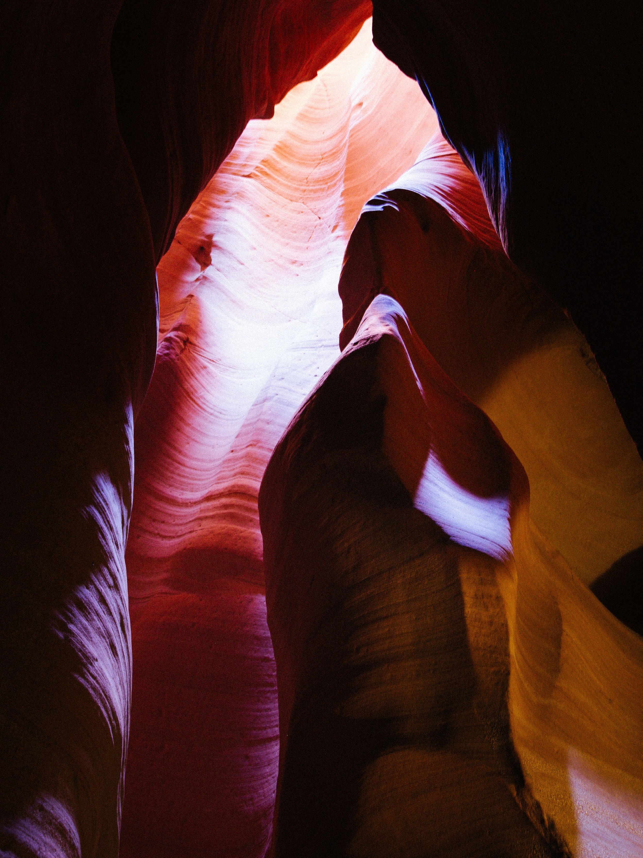 Inside a slot canyon with smooth, layered rock formations illuminated by natural light, showing vibrant red, orange, pink, purple, and yellow hues.