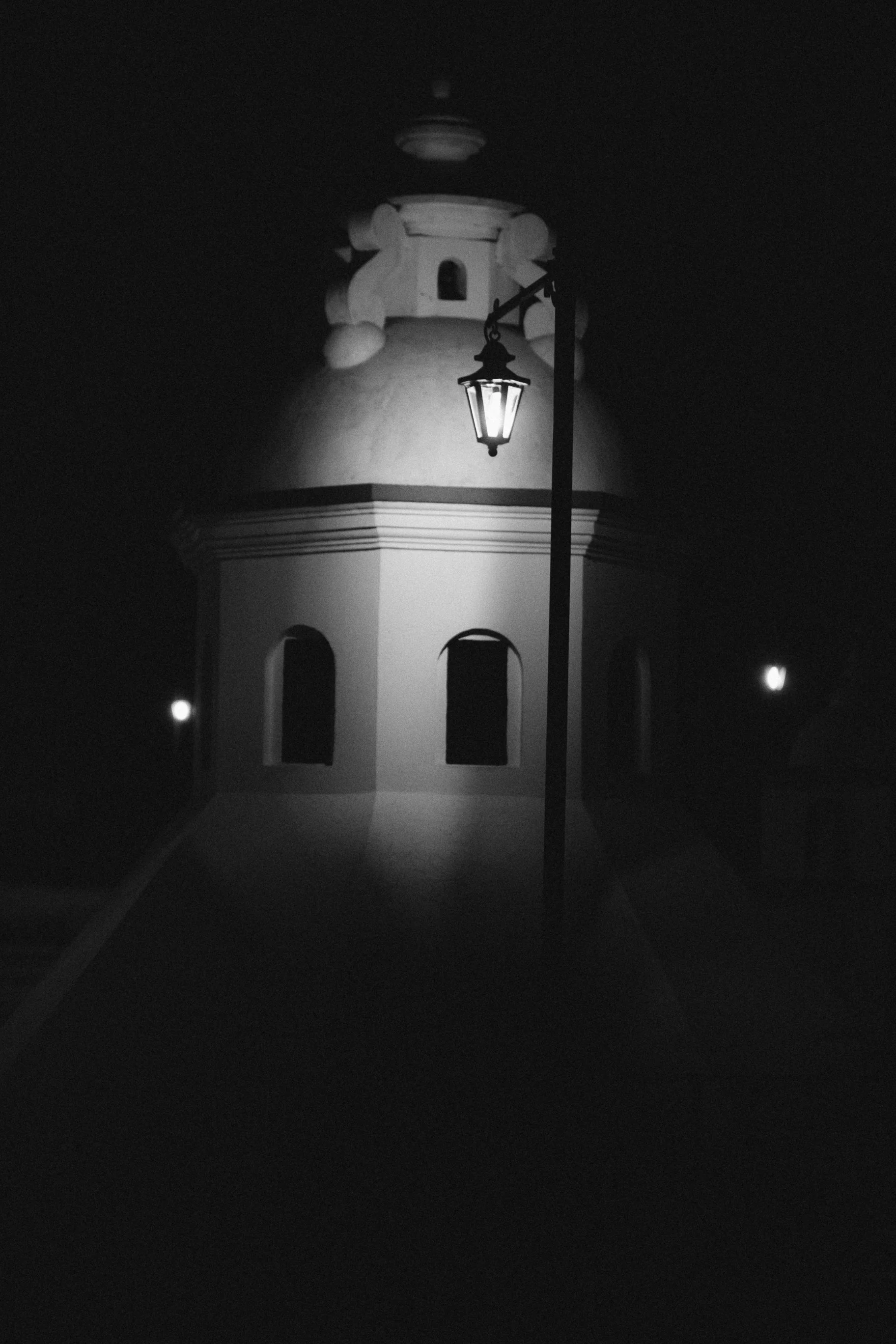 A black and white photo of a tower with a rounded top, illuminated by a streetlamp on a pole in front of it at night.