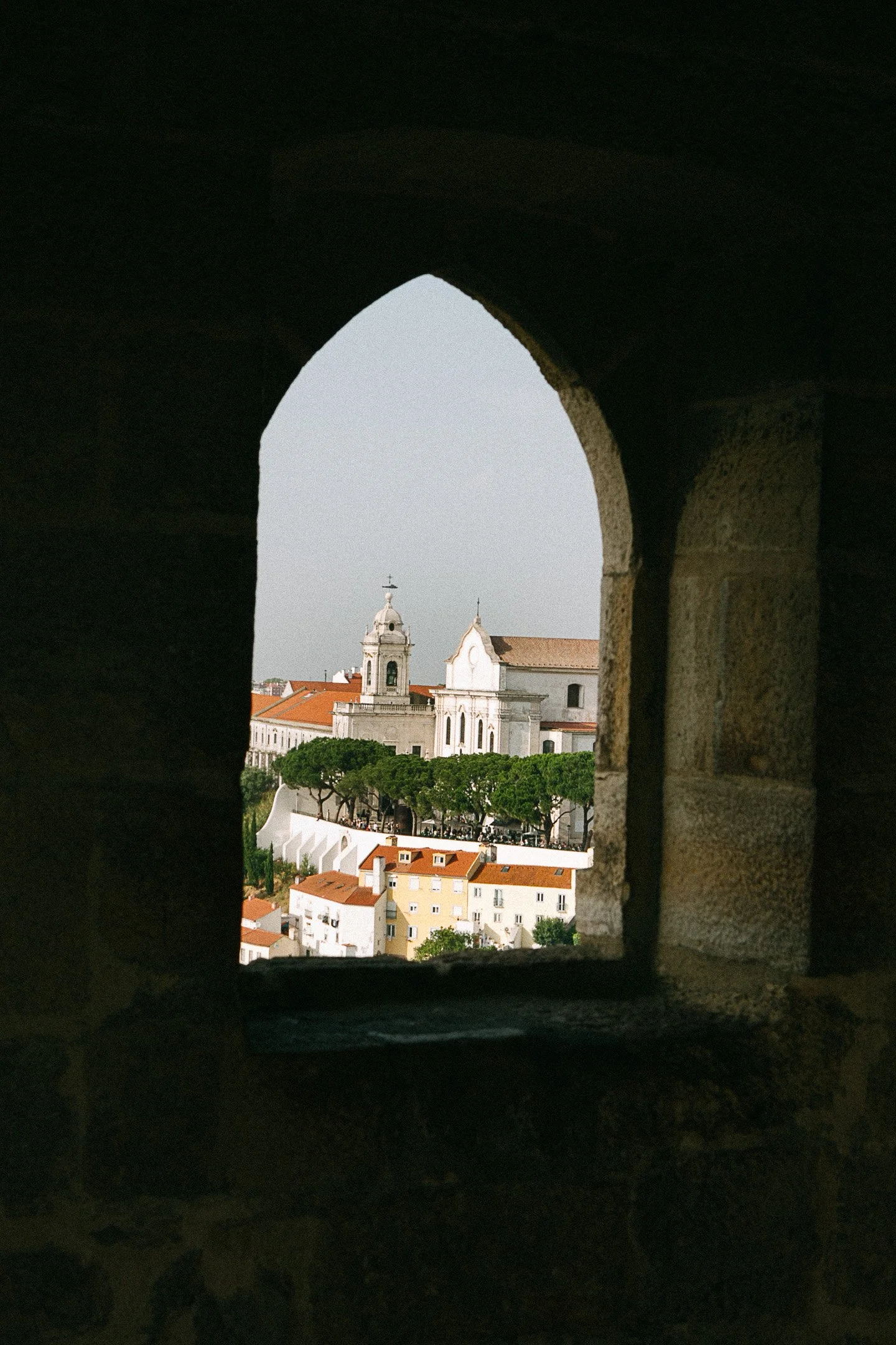 View of a European cityscape through a stone arched window, with historic buildings, a church with a bell tower, green trees, and red-tiled roofs.