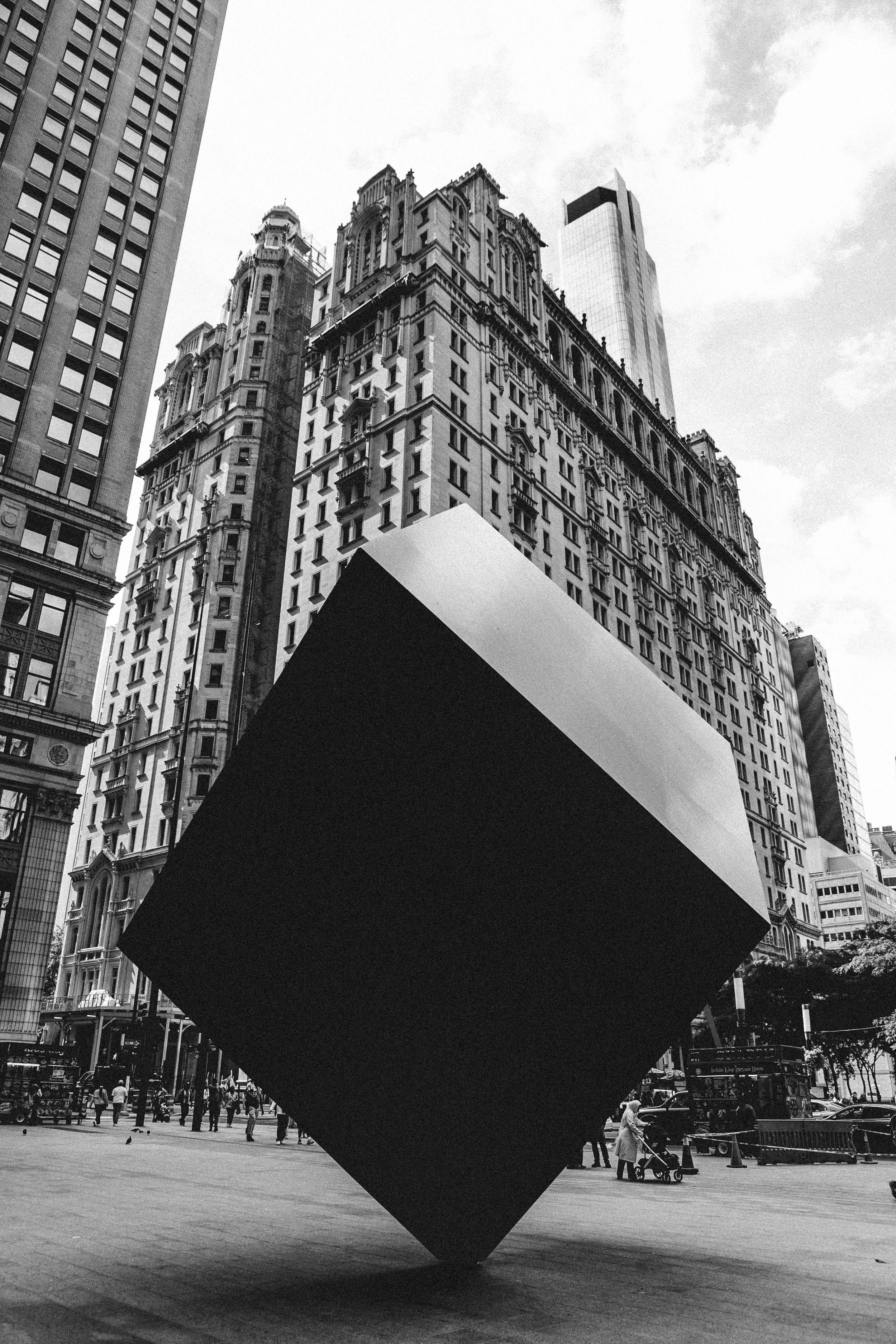 Black and white photo of a large geometric sculpture of a cube tilted on one corner in an urban plaza, with historic and modern tall buildings in the background, and pedestrians walking nearby.