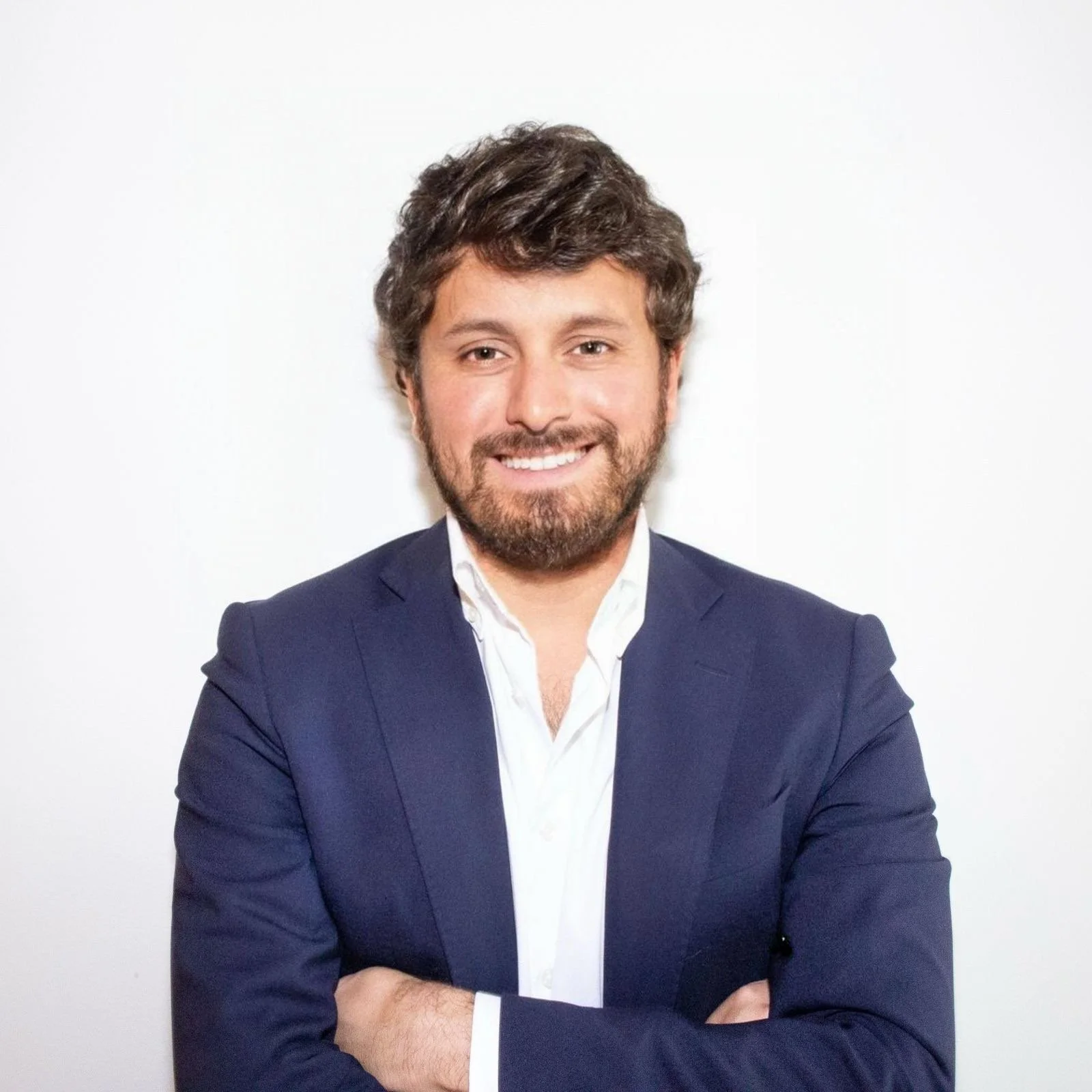 A man with brown curly hair and a beard, wearing a navy blue suit jacket and a white shirt, standing against a white background with his arms crossed and smiling.