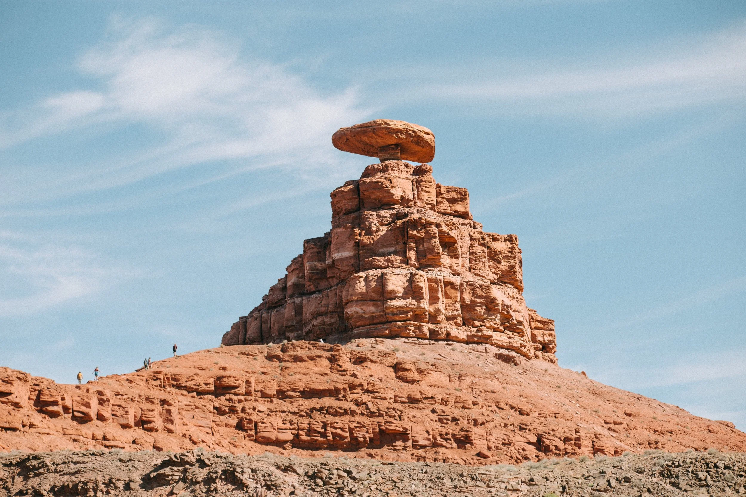 A large red rock formation with a flat, mushroom-shaped top in a desert landscape under a partly cloudy sky, with small figures of people walking nearby.