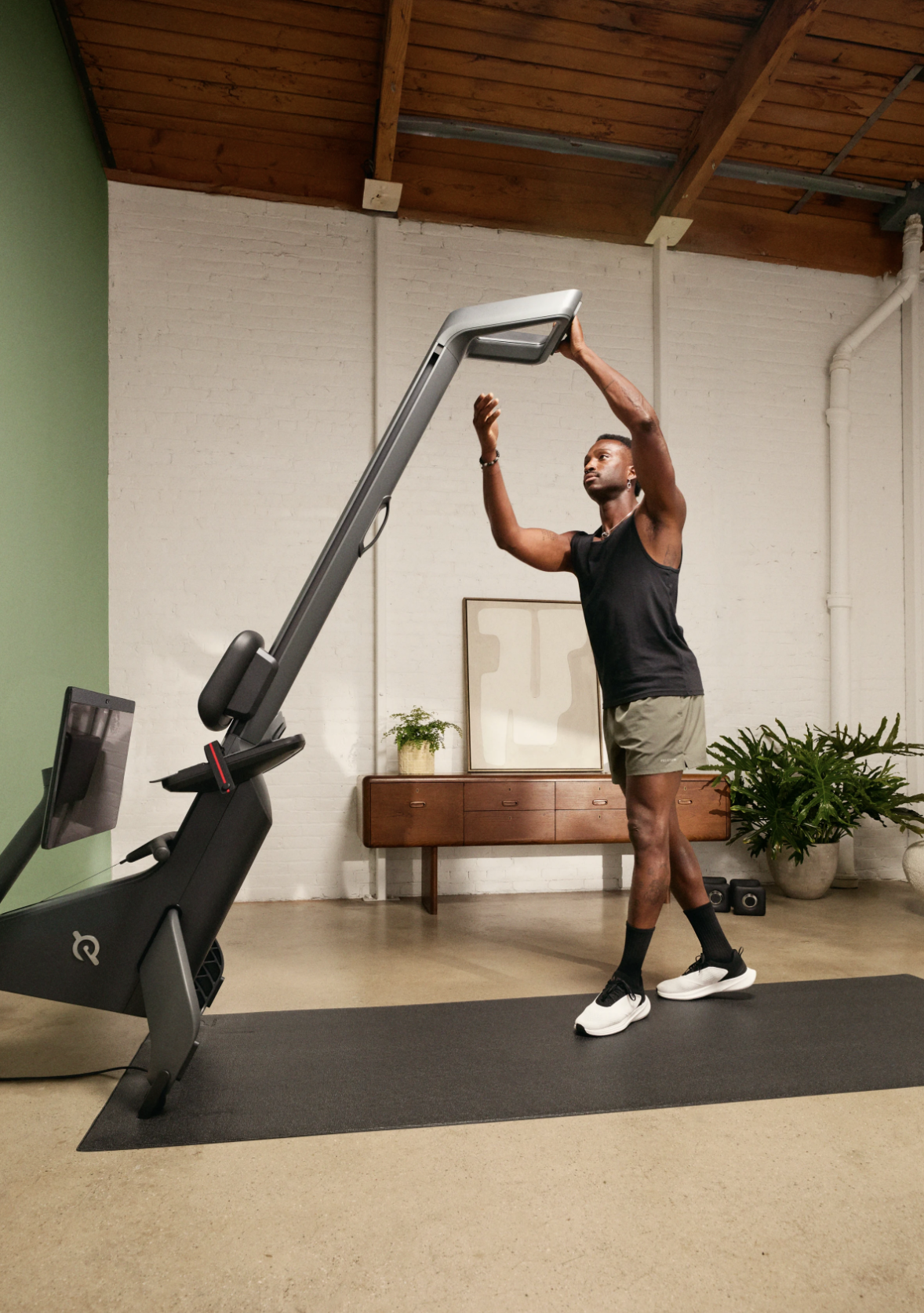 A man standing on a black exercise mat in a gym, stretching with his arms raised above his head, next to a stand-up exercise machine.