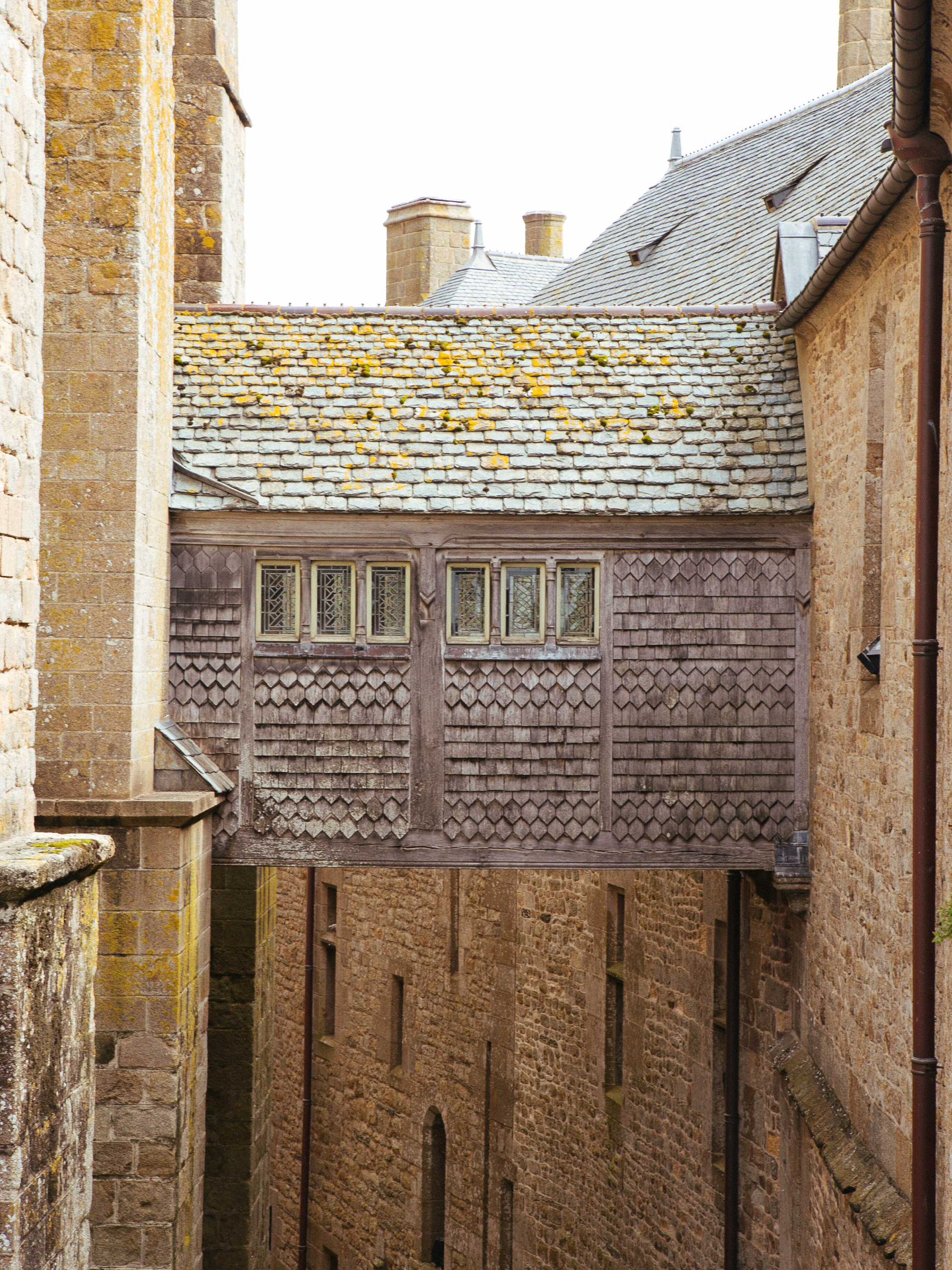 View of a historic stone building with a wooden bridge-like structure connecting two sections, featuring small leaded glass windows, moss-covered rooftops, and a narrow alley below.