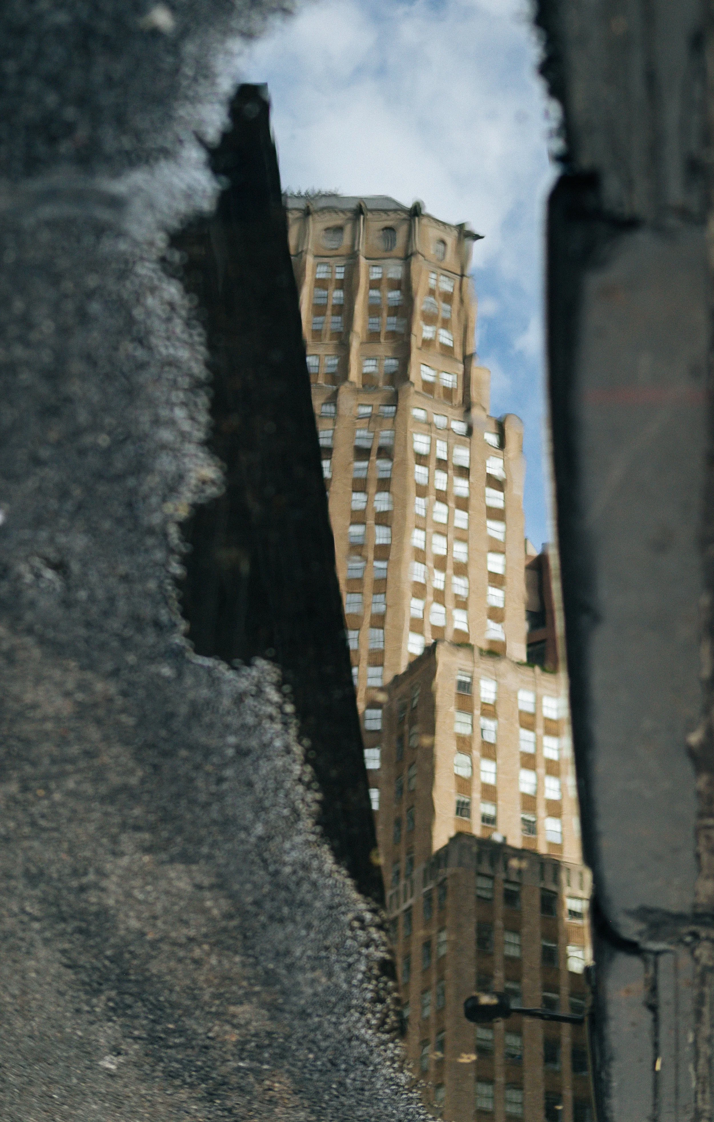 Reflected view of a tall, beige skyscraper with many windows, seen through a wet, narrow gap or crevice in a dark, textured surface, possibly a wall or structure, with a partly cloudy sky in the background.