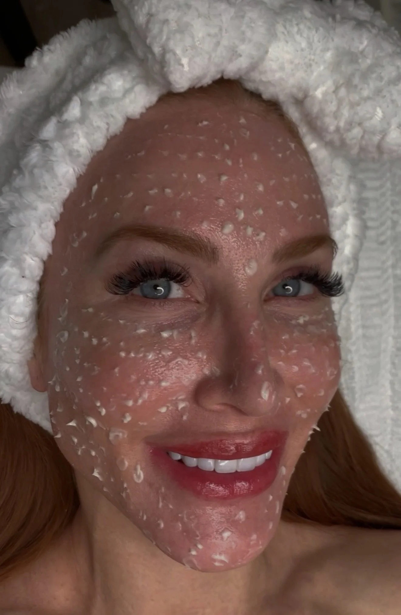 A woman with clear skin, blue eyes, long eyelashes, red hair, and red lipstick is lying down with a fluffy white towel wrapped around her head. She has a facial mask or treatment with white exfoliating particles on her face.