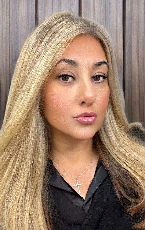 A woman with long, straight blonde hair, wearing a black top and silver cross necklace, standing in front of a wooden wall.