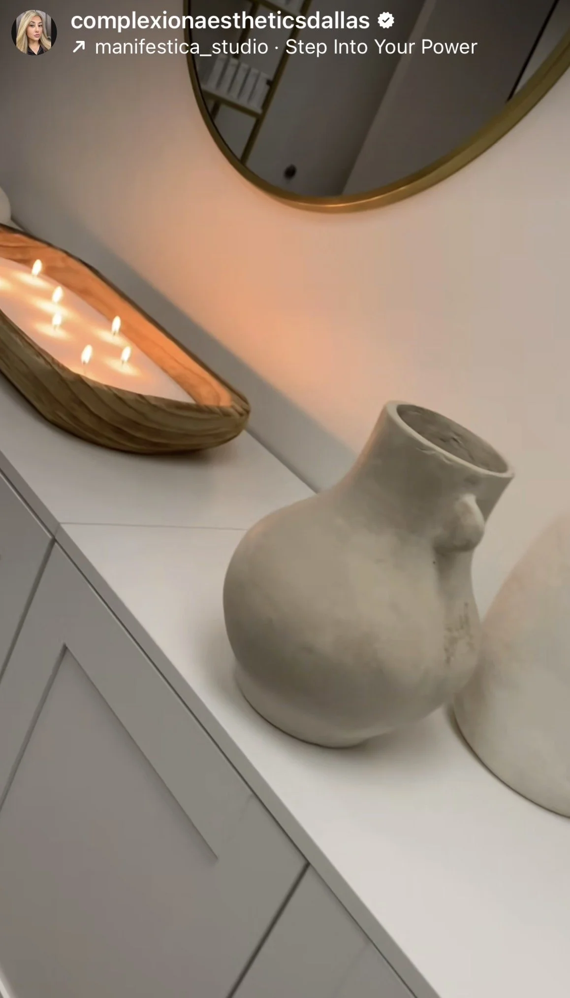 A white ceramic vase with an abstract, curvy shape on a white surface, with a wooden tray with candles and a round mirror on the wall above.