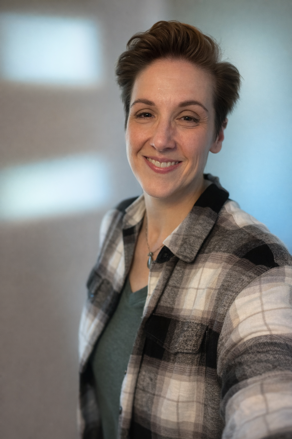 A woman taking a selfie indoors, smiling, with short brown hair, wearing a plaid shirt over a green top.
