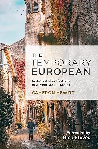 Book cover titled 'The Temporary European: Lessons and Confessions of a Professional Traveler' by Cameron Hewitt, with a subtitle 'Foreword by Rick Steves.' The background features a European street with stone buildings and cobblestone pavement. Book