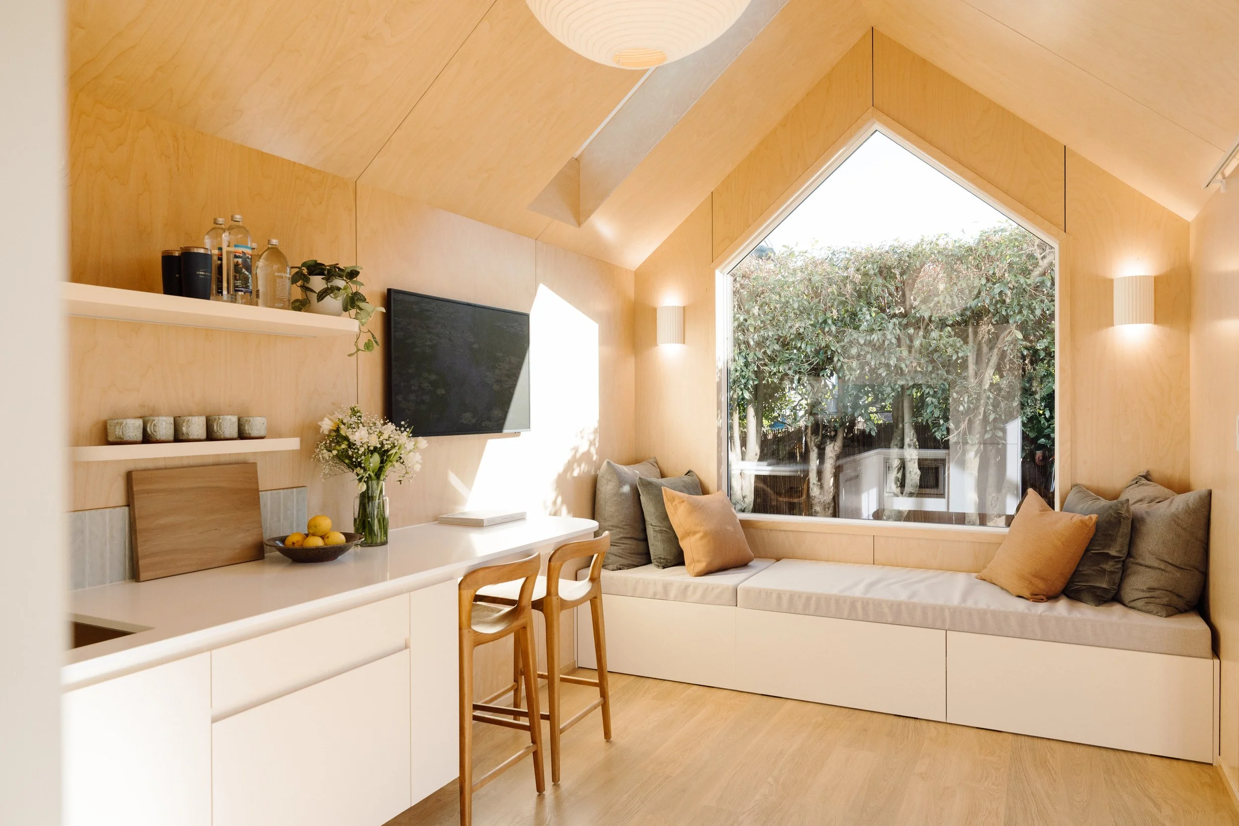 A cozy living space with wooden walls and ceiling, featuring a large triangular window showing green trees outside. The room has a built-in cushioned seating area with pillows, a wall-mounted television, a white shelf with bottles and a plant, and a small dining area with a vase of flowers and a bowl of lemons.