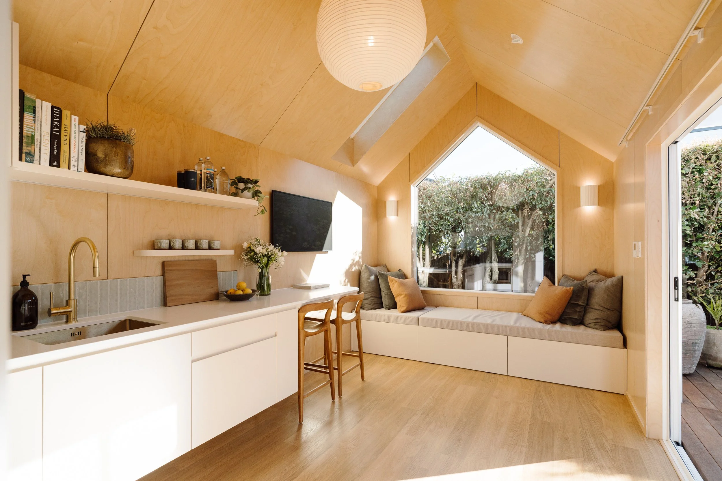 Interior view of a small, cozy living space with birch low VOC plywood walls, a built-in daybed that doubles as an extra queen size bed, a full kitchen, TV, WIFI and large beautiful windows letting loads of sunlight in.