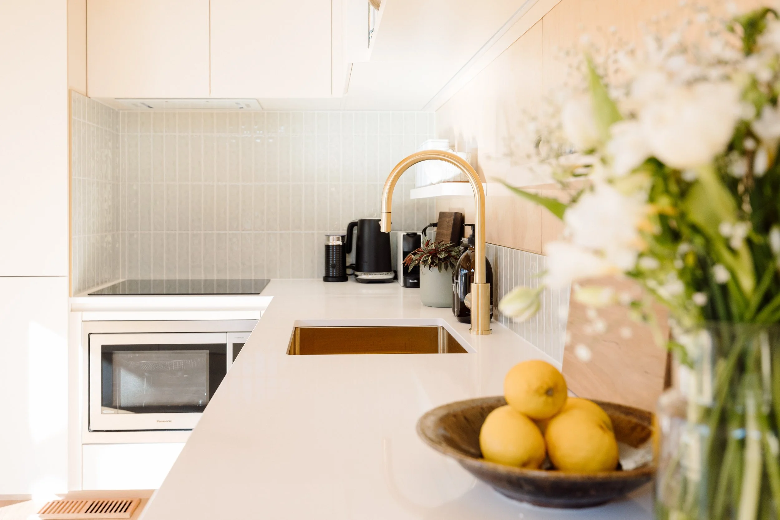 A modern kitchen with white countertops, a brass faucet, and a bowl of lemons, with flowers partially visible in the foreground.