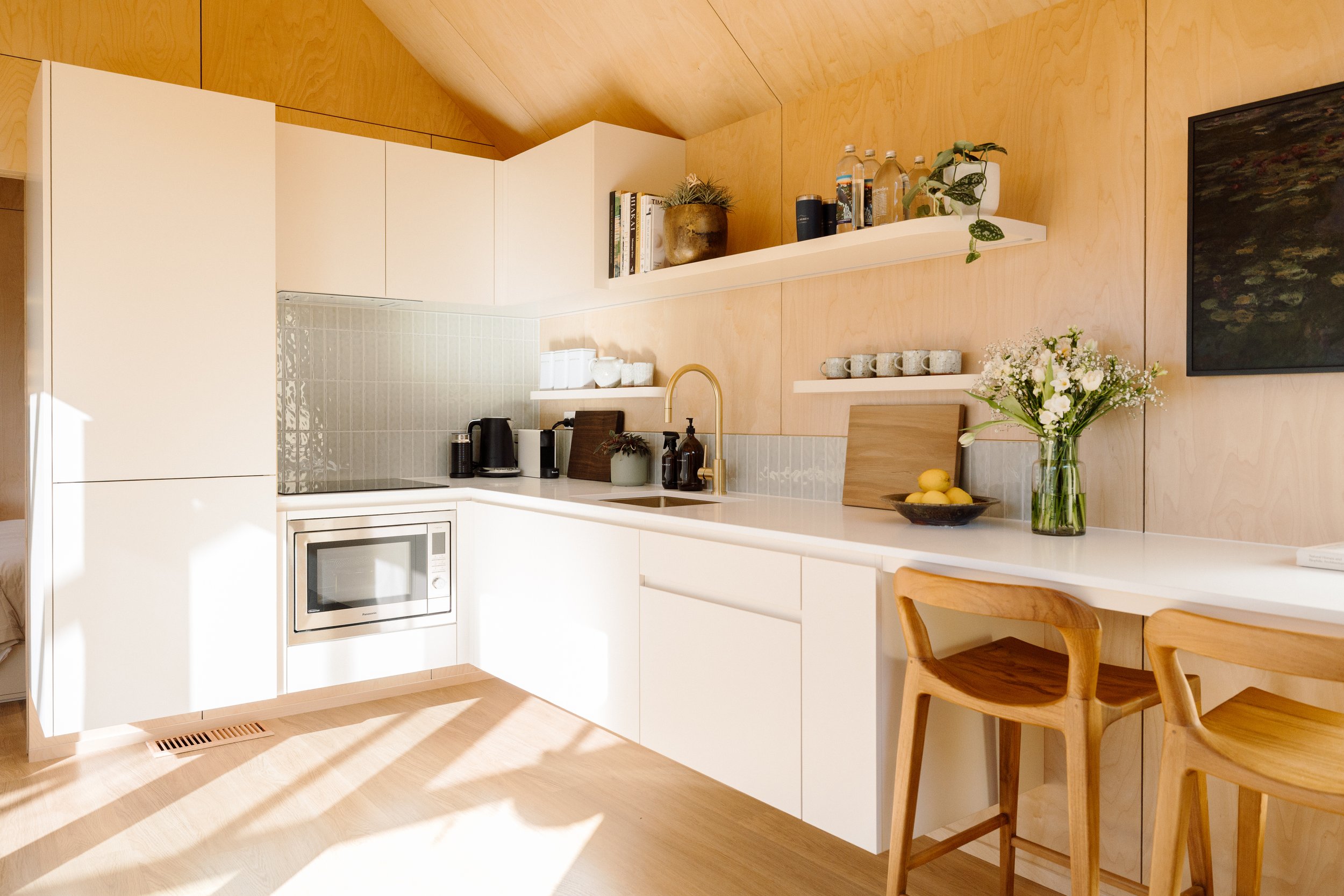 A modern kitchen with white cabinetry, a built-in microwave, a brass faucet, open shelves with books, plants, water bottles, a bowl of lemons, and a flower vase with white flowers.